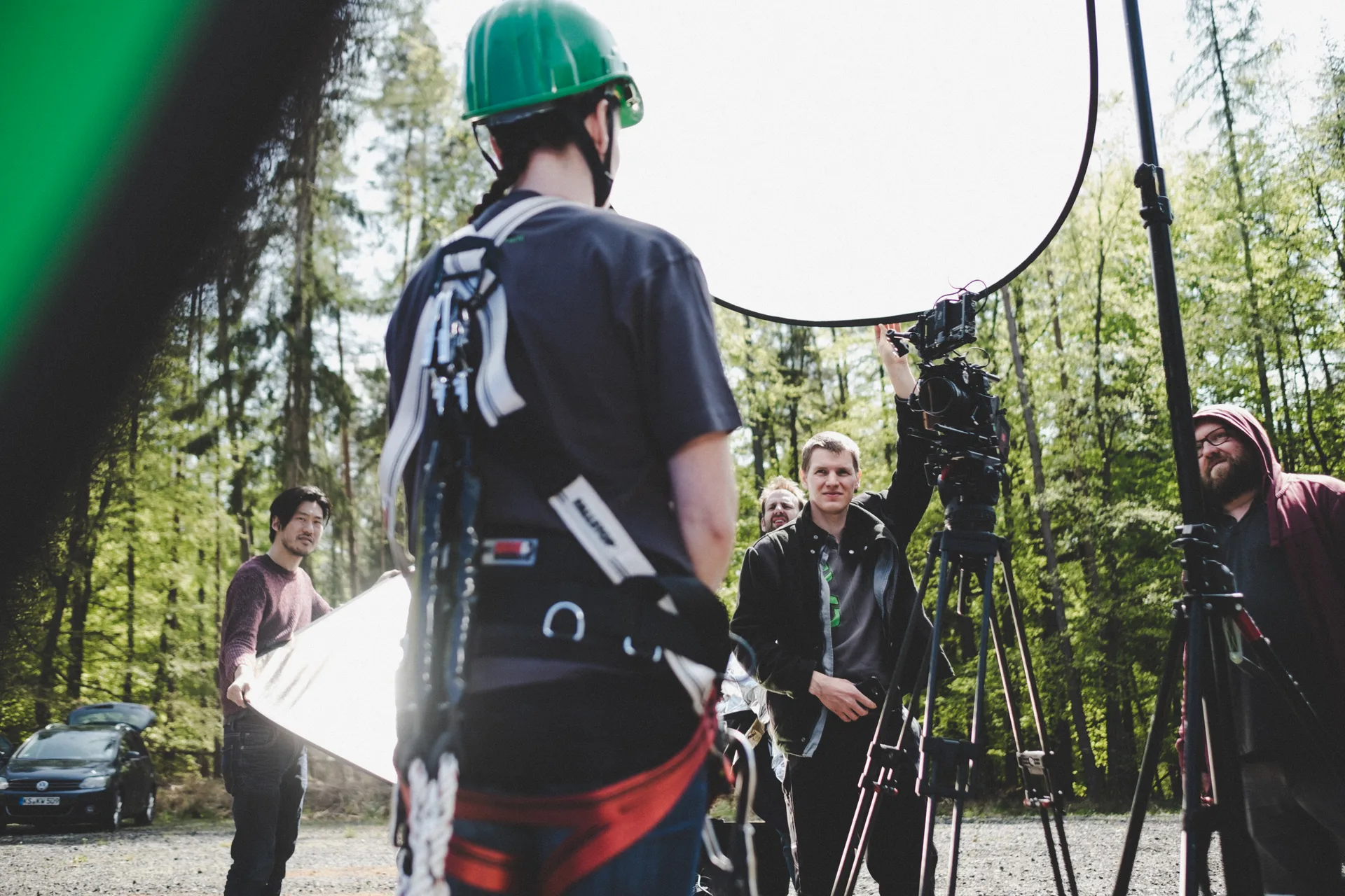 Film crew on location in a forest, with a camera on a tripod, reflector, and people in workwear.