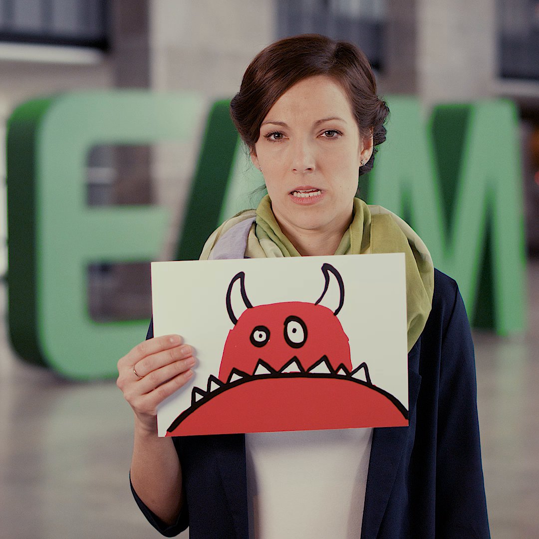 A woman holds a picture of a red monster, with large green EAM letters visible in the background.