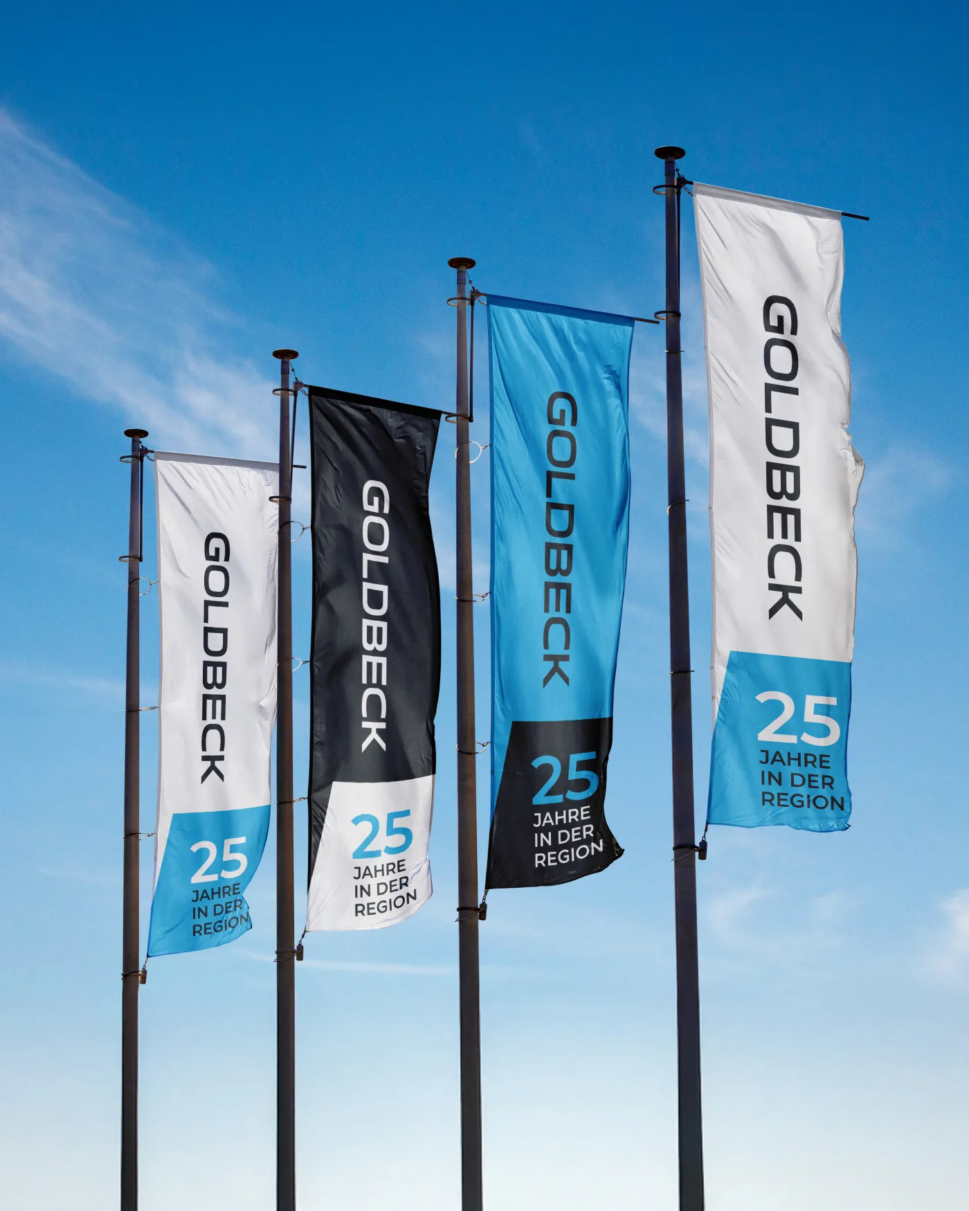 Four Goldbeck flags in black, white, and turquoise, bearing the inscription '25 years in the region', waving against a blue sky.