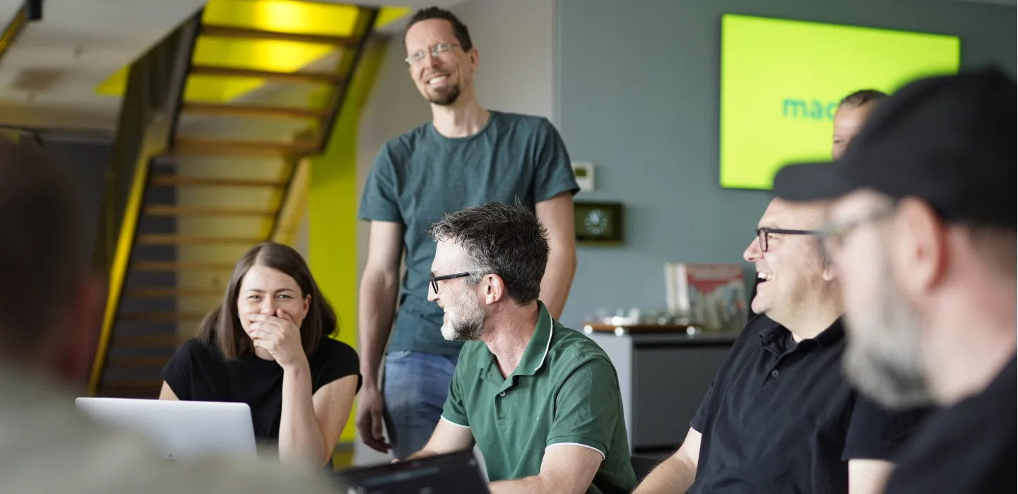 A team of six people laughing and talking in an office with a yellow-green staircase and a screen displaying the "machbar" logo.