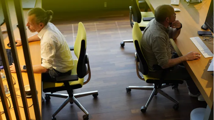 Two men sit at wooden desks, working on computers; the one on the left has a bun and a white shirt, the one on the right has a beard and a grey shirt.