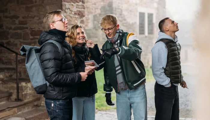 Four young people, three men and one woman, are standing outside and looking around, while one of them points at something.