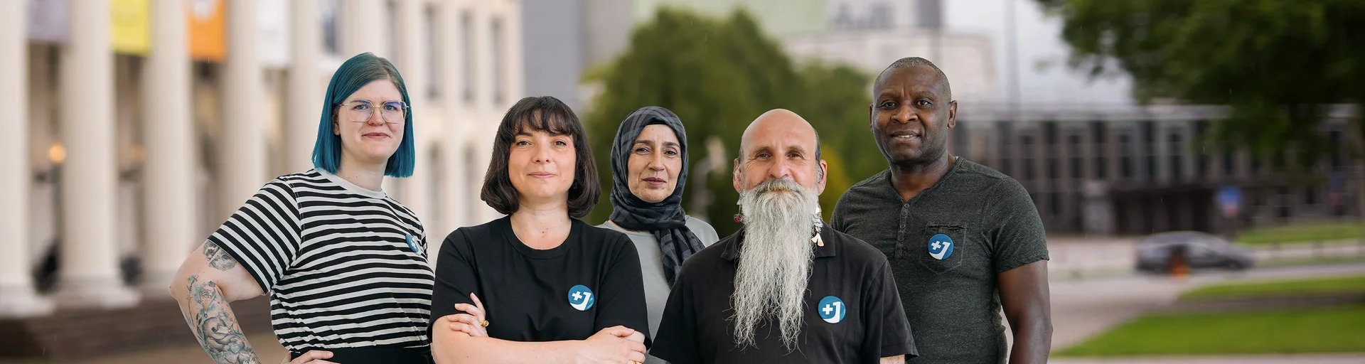 Fünf Personen unterschiedlichen Alters und Herkunft, darunter eine Frau mit blauen Haaren und eine Frau mit Kopftuch, tragen T-Shirts mit dem "+1"-Pin der Kampagne "Backup – Gegen Hass, gegen Hetze".