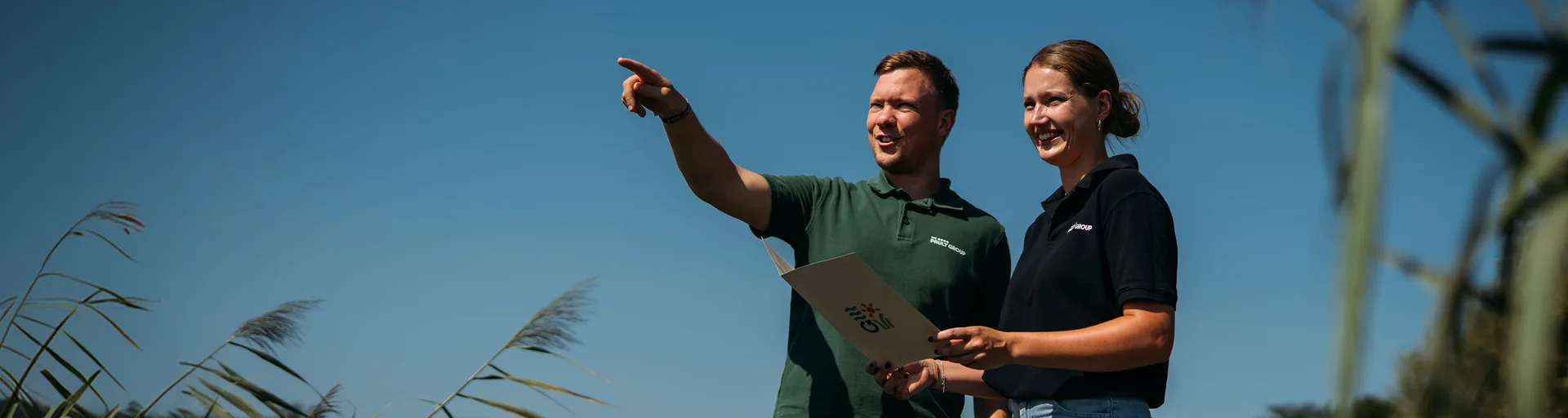 Two people in workwear stand against a blue sky and reeds; the man points, the woman holds a document with the 'GII' logo.