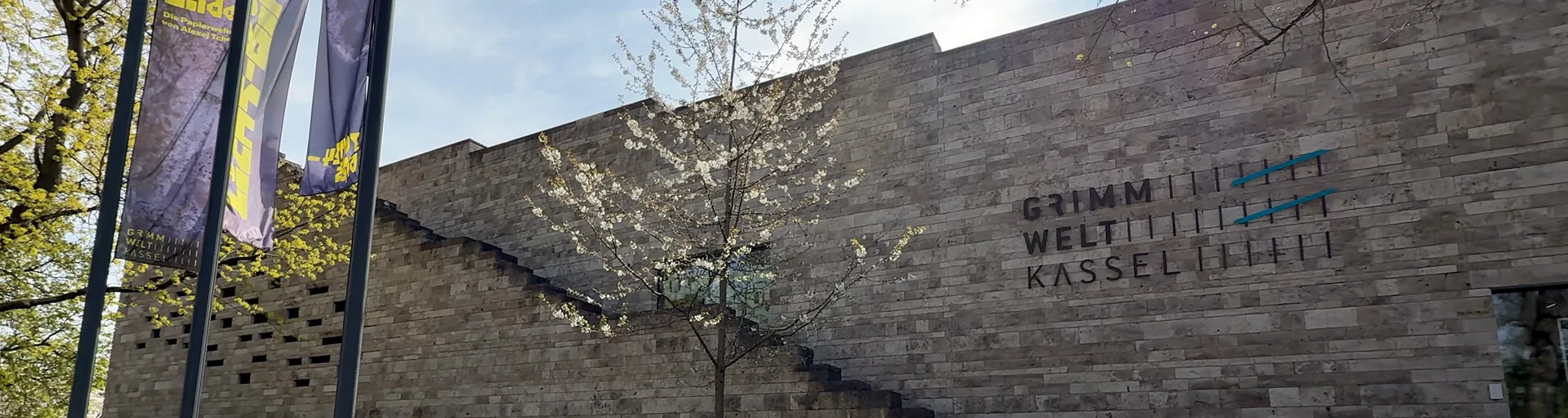 Die Fassade der Grimmwelt Kassel mit dem Schriftzug "GRIMMWELT KASSEL" und einem blühenden Baum im Vordergrund.