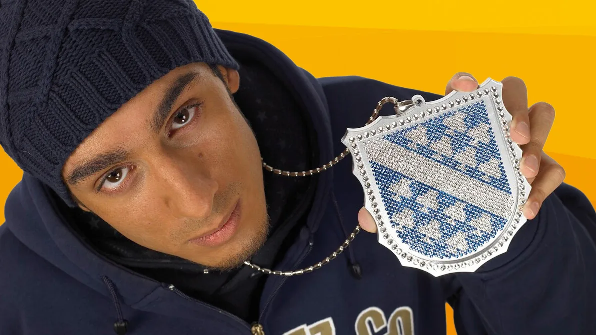 A young man in a dark blue knitted hat and hoodie holds a glittering chain with the Kassel coat of arms up to the camera.